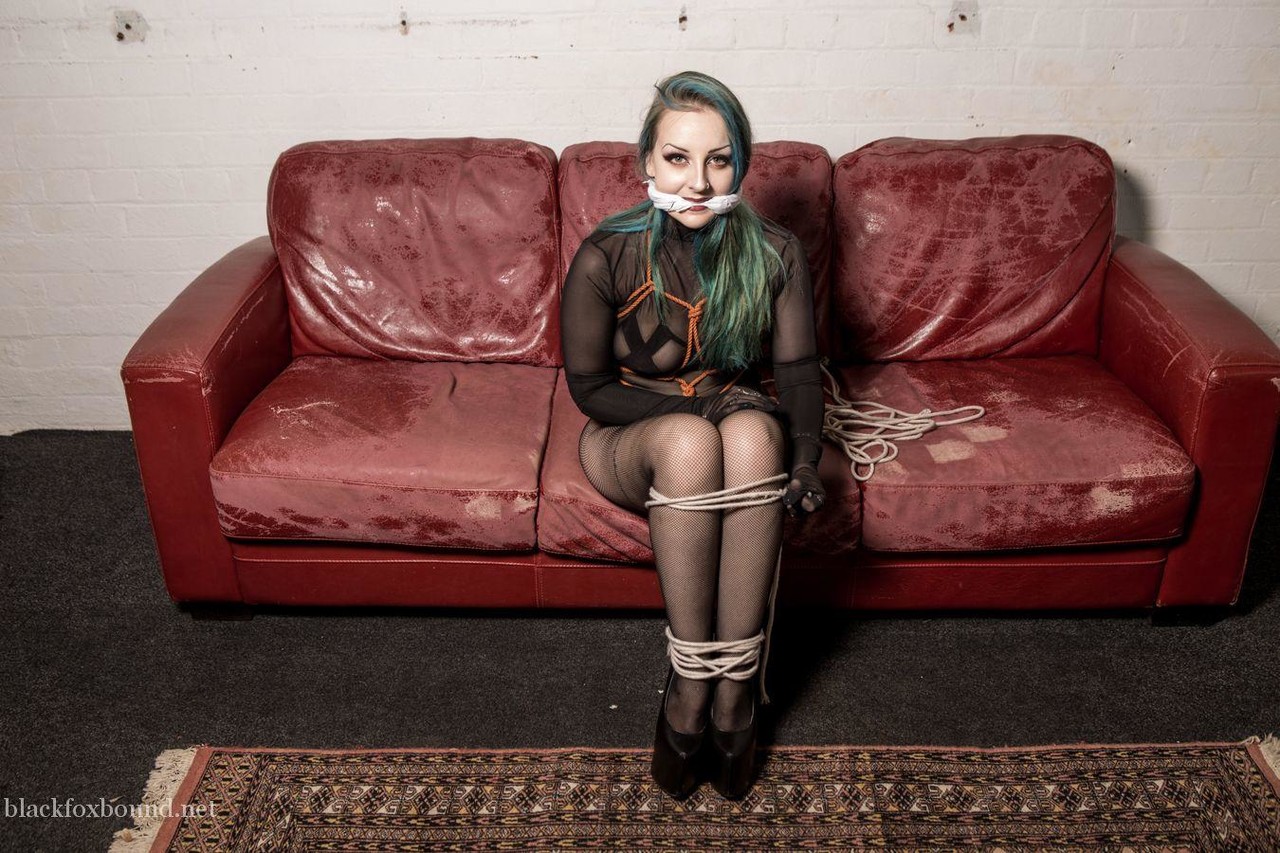 A white girl with dyed hair is gagged and hogtied on a Chesterfield.