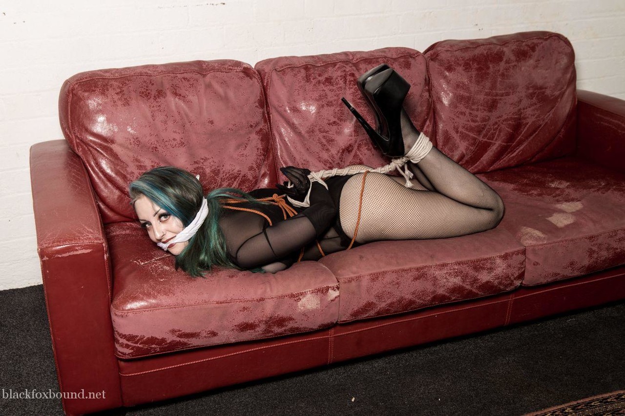 A white girl with dyed hair is gagged and hogtied on a Chesterfield.