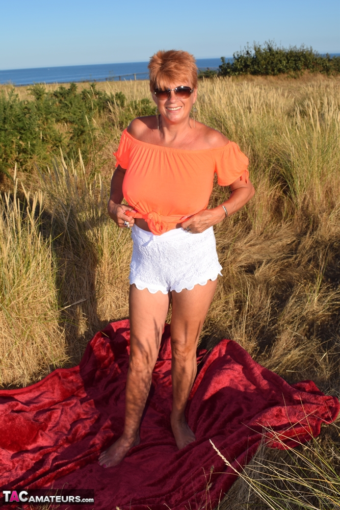 Dimonty, a fit elderly woman who is also tall, exposes large natural wonders on a blanket that overlooks the ocean.