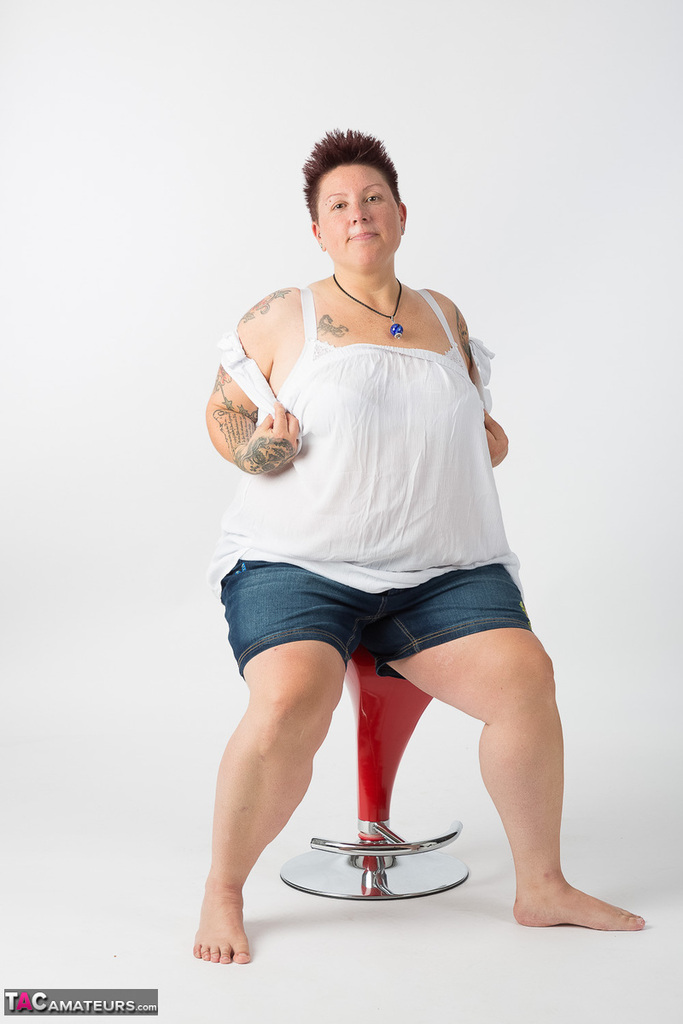 After getting completely naked, a tattooed amateur gets up and sits on top of a stool.