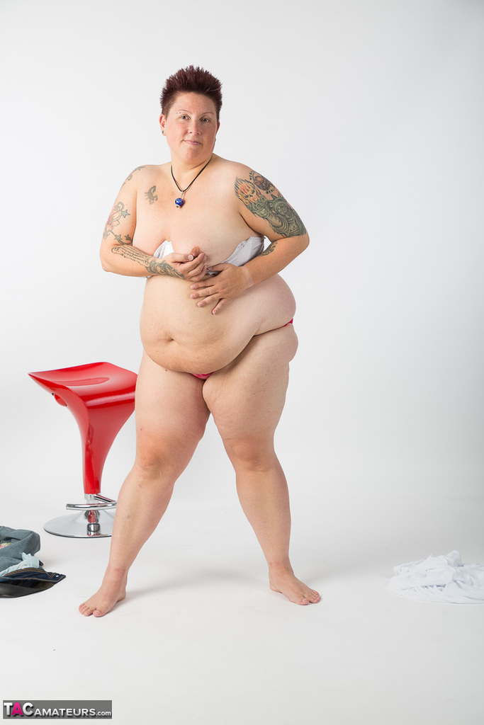 The naked tattoo girl, who is an older woman than the average, sits on a stool.