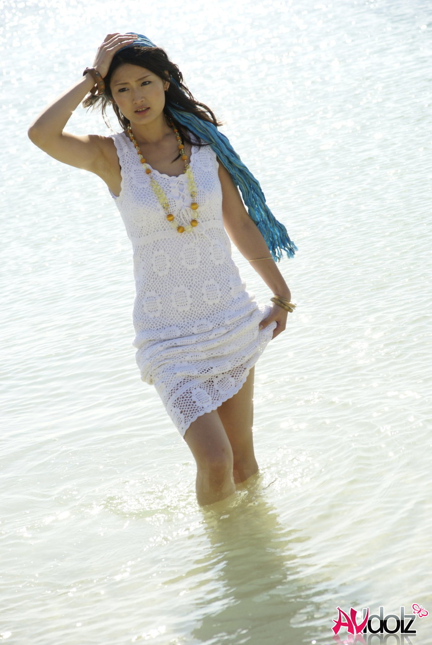 Donning a white dress, an Asian girl walks into the ocean with her knees raised.