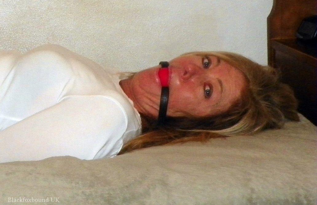 Restrained: A blond MILF is ball gagged and restrained on a bed in black miniskirt form.