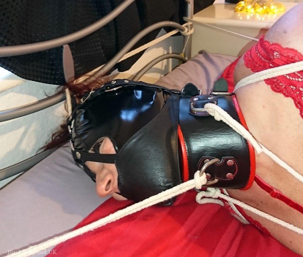 A white girl with a collar is tied down to a bed in red lingerie and a latex hood.