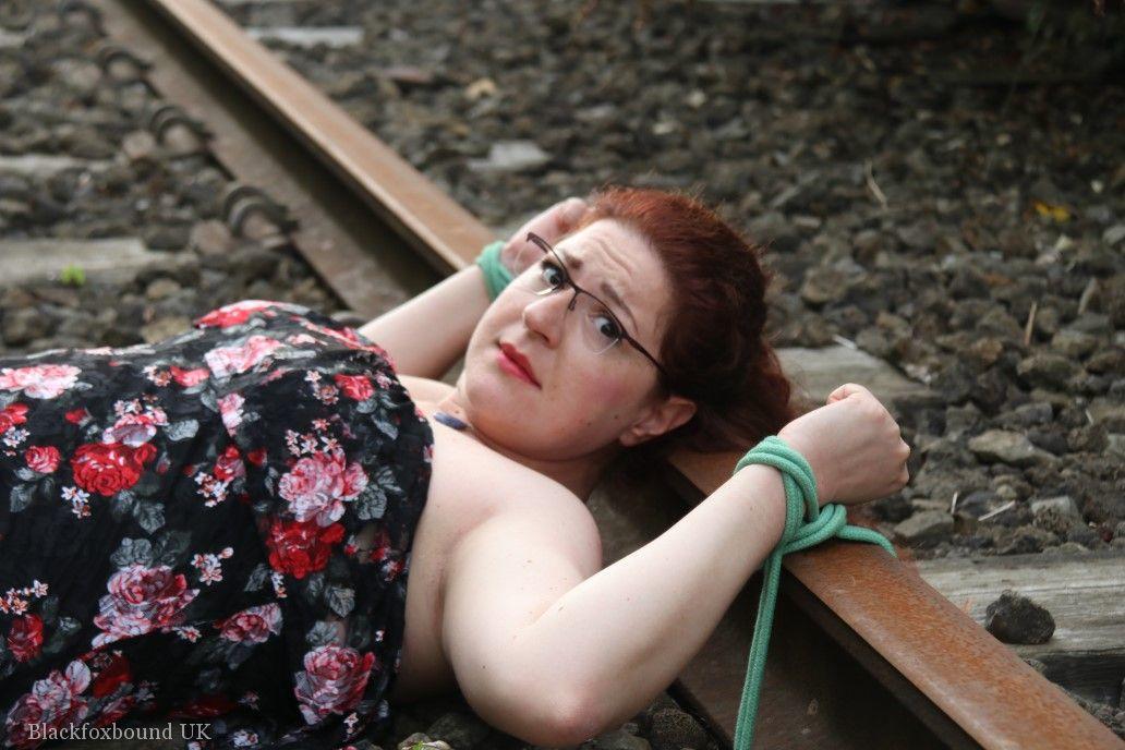 Wearing a dress and glasses, the natural redhead is tied to train tracks.