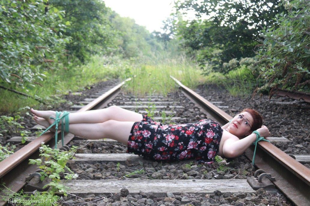 The natural redhead is wearing a dress and glasses and has been tied down to the train tracks.