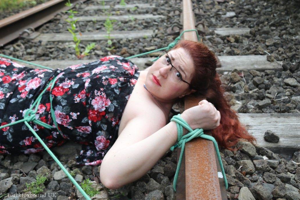 Wearing a dress and glasses, the natural redhead is tied to train tracks.