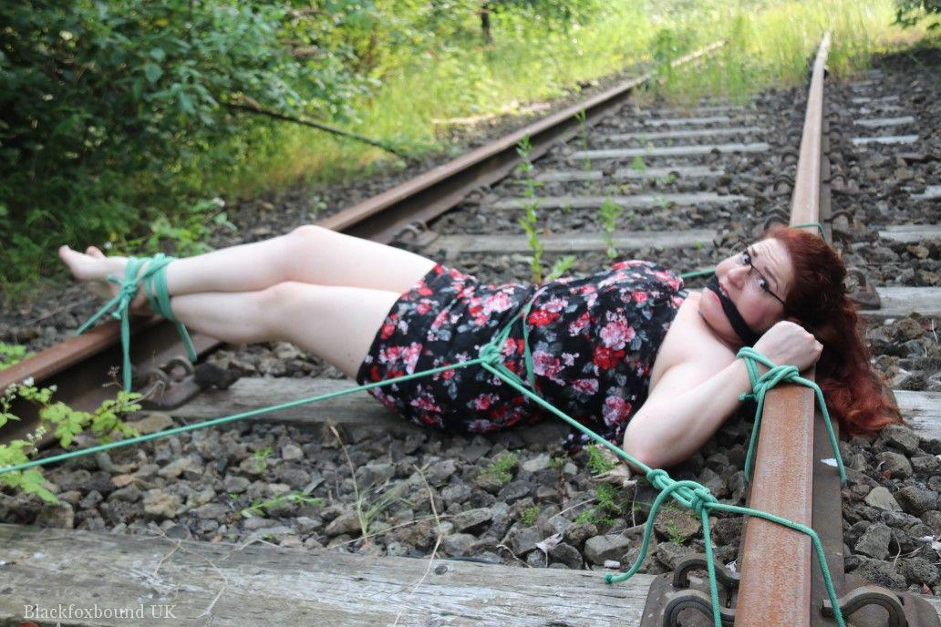 The natural redhead is wearing a dress and glasses and has been tied down to the train tracks.