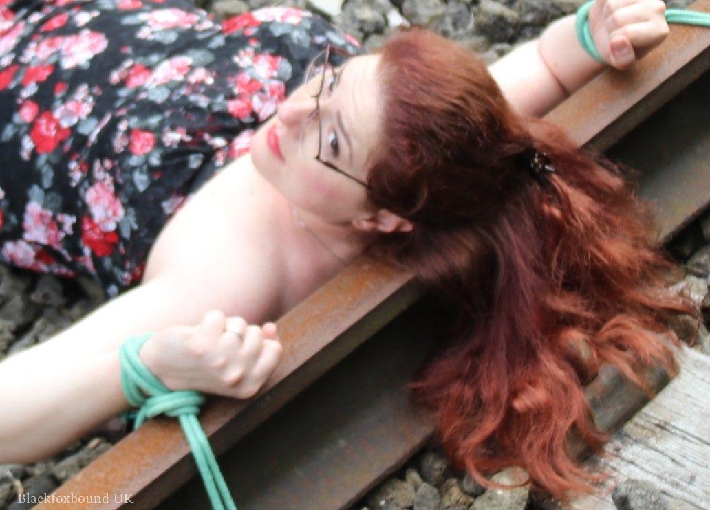 Wearing a dress and glasses, the natural redhead is tied to train tracks.