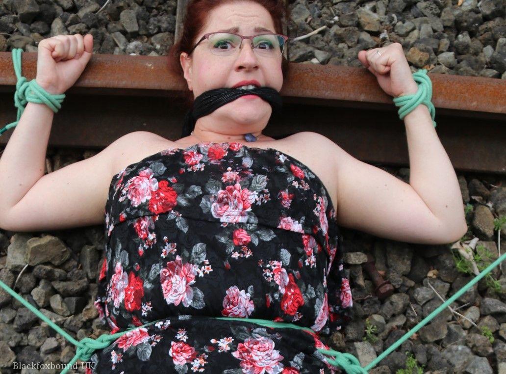 The natural redhead is wearing a dress and glasses and has been tied down to the train tracks.