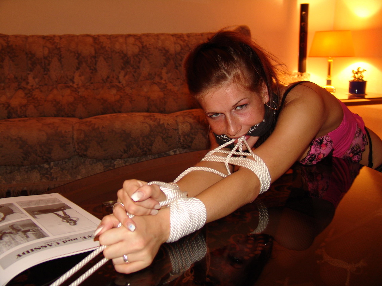 White girl sporting cleave gag while restrained with rope draped over coffee table.