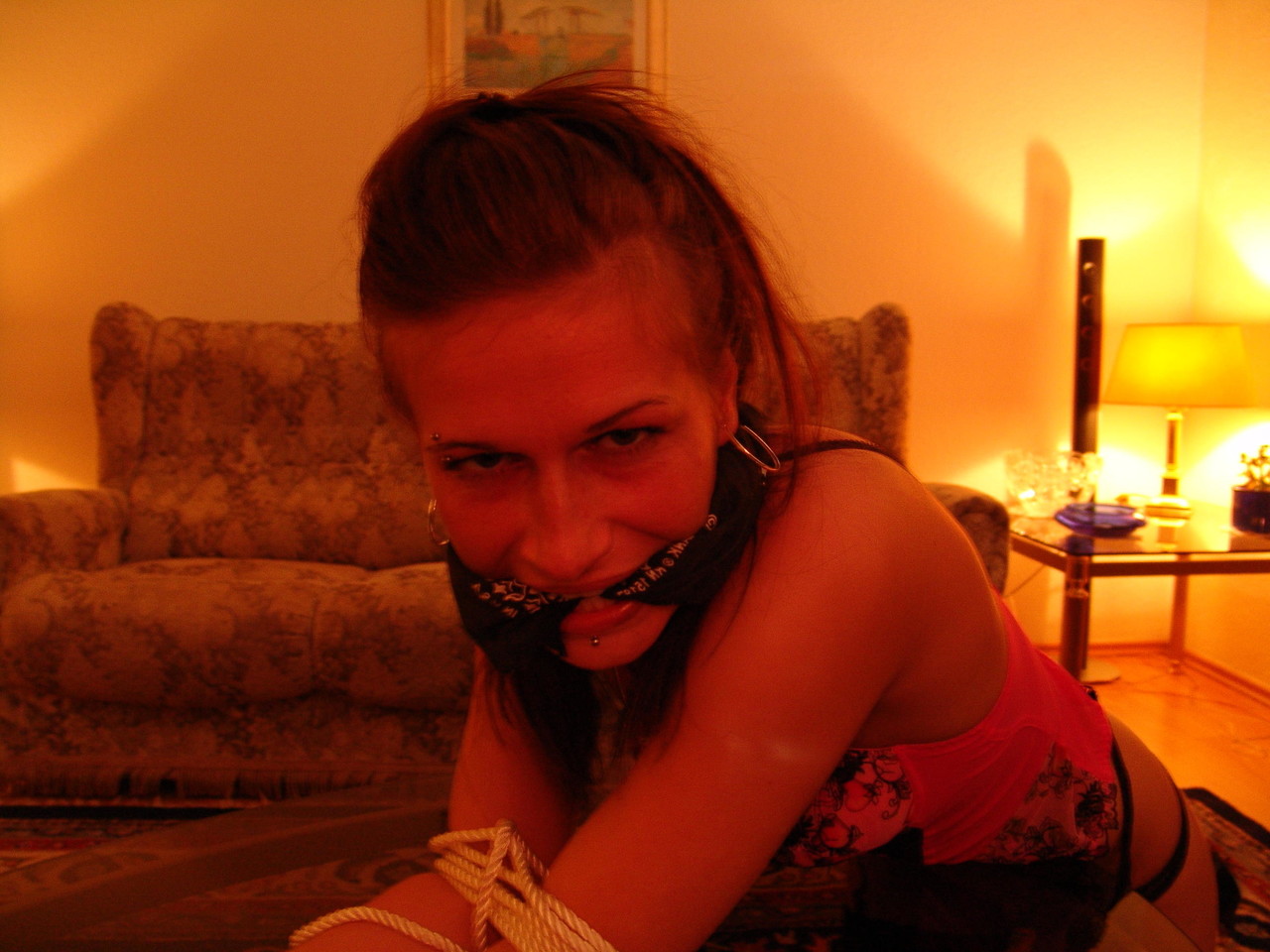 A white girl exhibits a cleave gag while being restrained with rope over a coffee table.