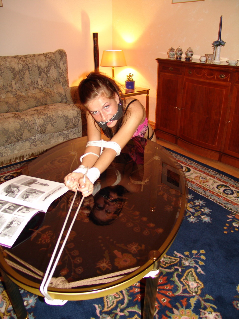 A coffee table is adorned with an exposed rope and a white girl sporting a cleave gag.