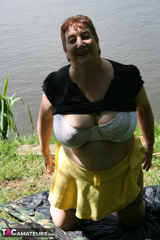 An older redhead named Kinky Carol puts on a bra and layers of clothing near the water.