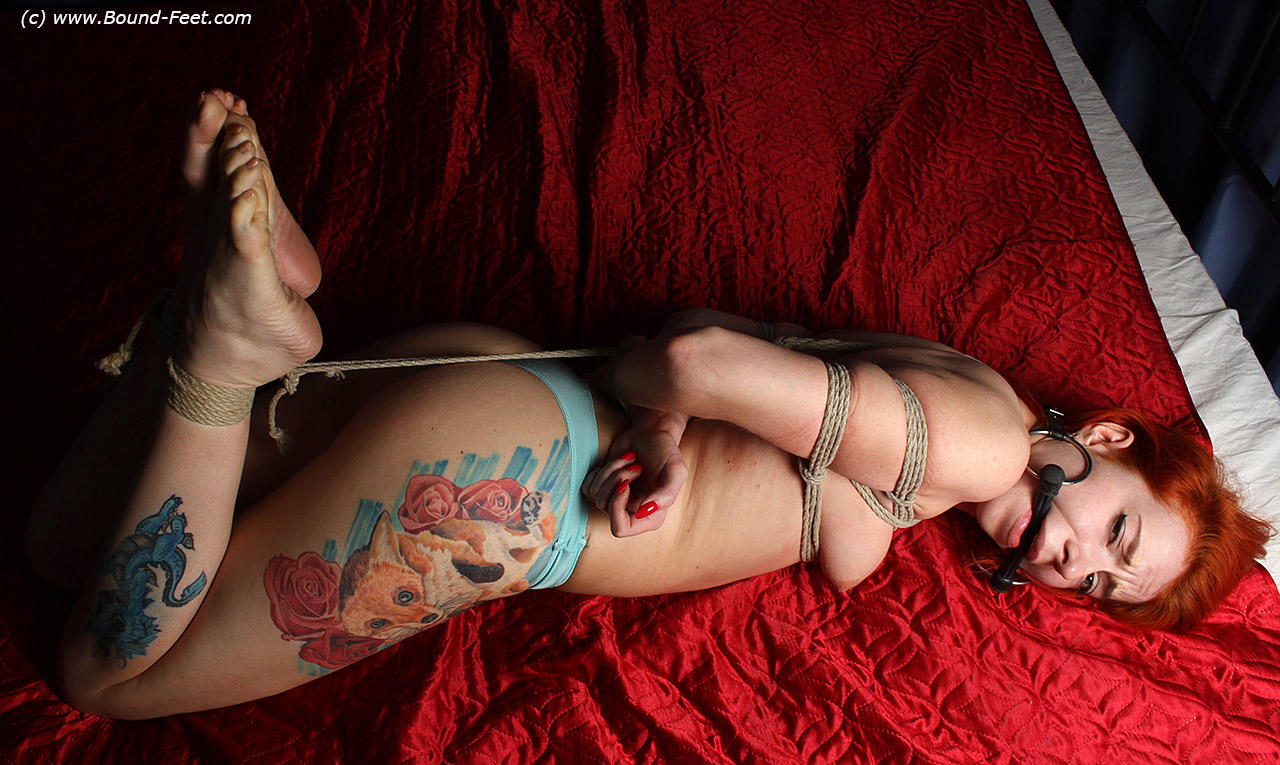 With a horse biting him, a tattooed redhead is restrained on a bed with rope.