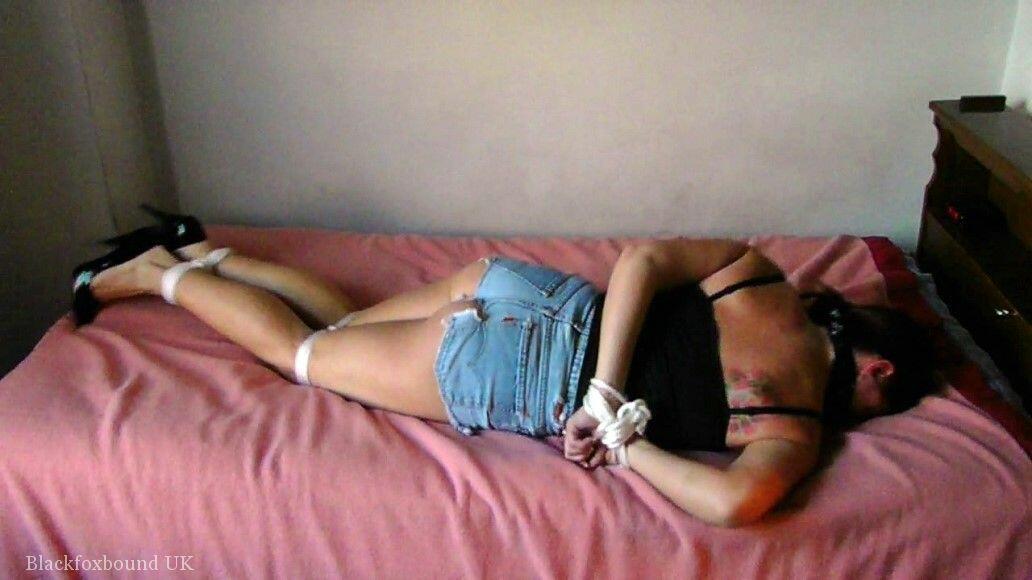 The brunette female is tying up and gagged while lying on a bed, with her left side facing down.