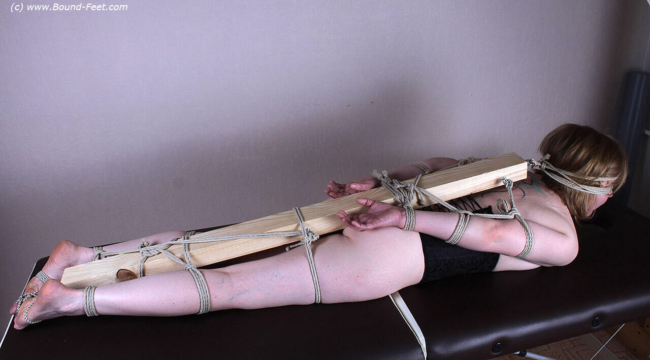 A 2 x 4 is secured behind the back of a Caucasian female, with her feet, arms, and legs connected.