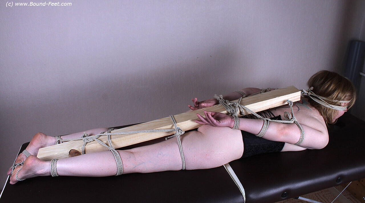 Behind the female figure, a 2'x 4 is being tied to her feet, arms, and legs.