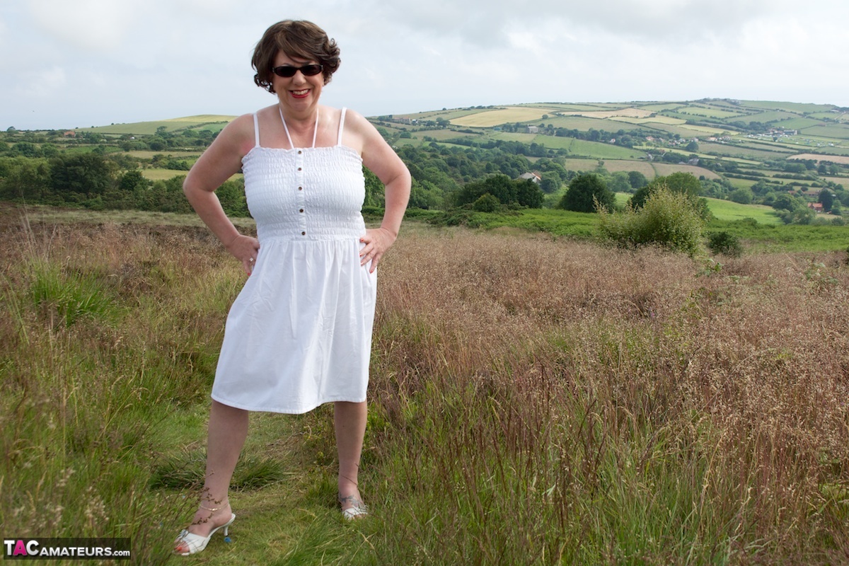 In a field, the elderly lady Dirty Doctor sits on top of a blanket and goes naked.