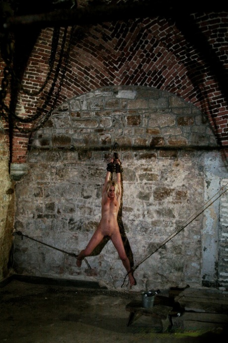 A naked blonde named Mirela is chained to a cellar wall while being whipped by captors.