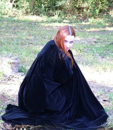 Amber Lily, a redhead amateur, models herself naked in a forest while wearing a black cape.