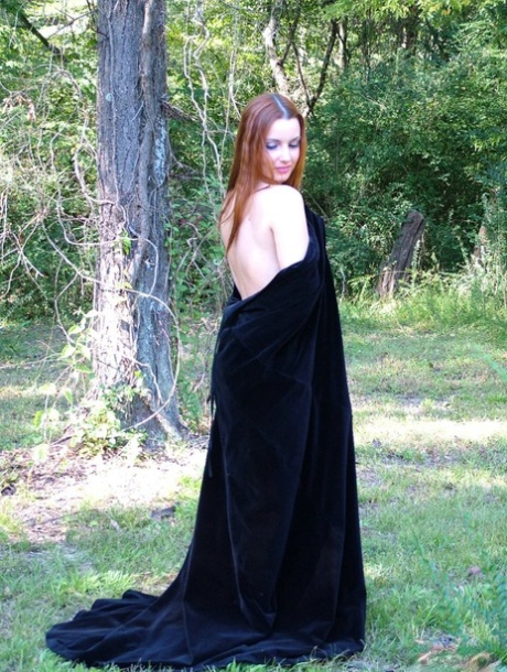 In a forest, Amber Lily, a redhead amateur, models herself naked in her black cape.