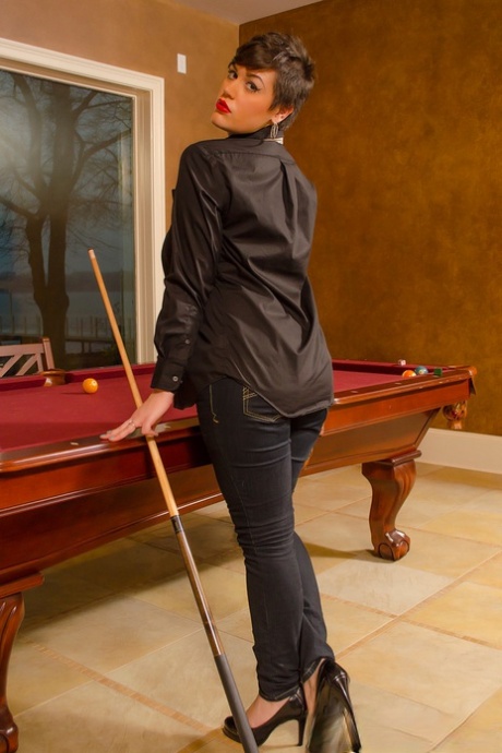 In front of a pool table, Annalise, a thin and petite woman with short hair, exposes her large breasts and buttocks.