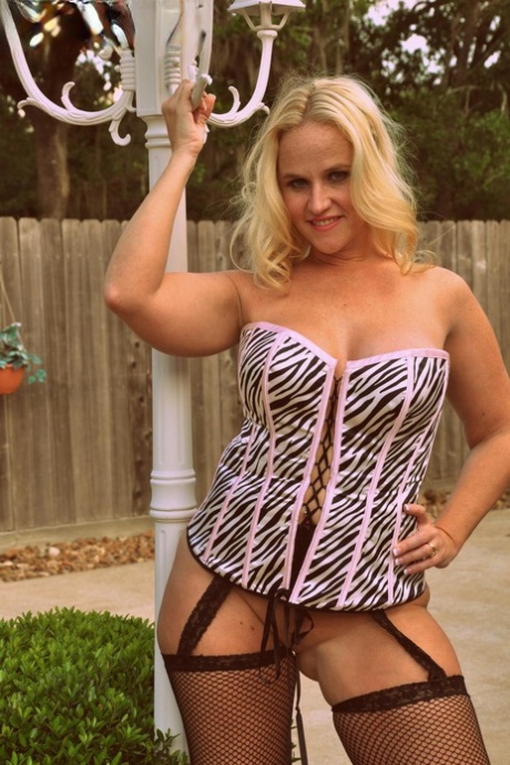 Dee Siren, a blonde amateur, models herself wearing a laced corset and mesh hosiery in a backyard setting.