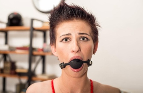 The girl with short hair is attired in a ballgag and jeans, tied up without a top.