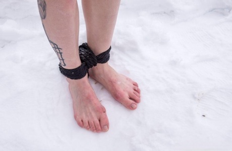 In the snow, a girl with tattooed feet is forced to kneel and stand barefoot due to her condition.