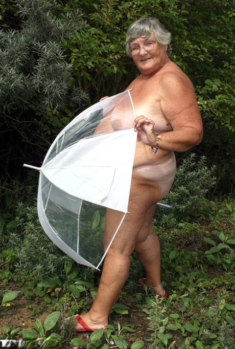 An umbrella is held by Obese OMa Grandma Libby while she poses unclothed by fir trees.