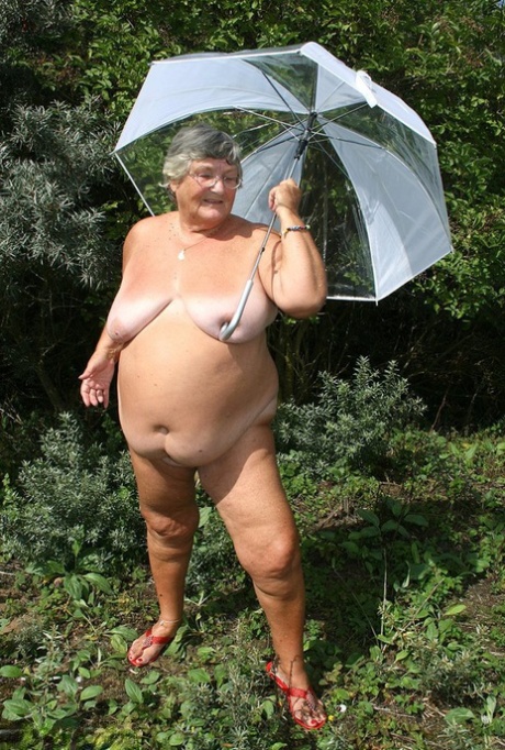 Grandma Libby is seen holding an umbrella while unclothed by pine trees.