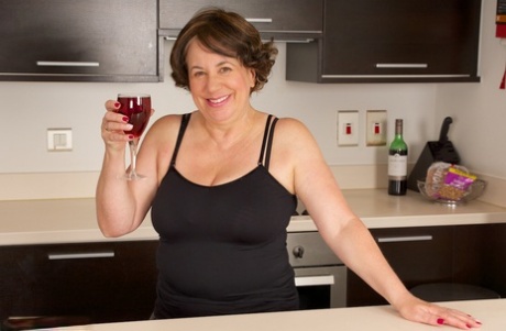 Before revealing her large breasts on a kitchen counter, the elderly lady raises herself a glass of champagne.