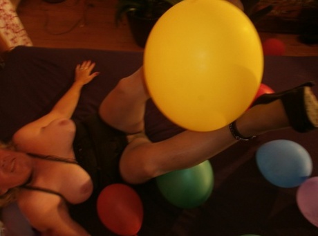 The old woman named Caro displays her breasts in pantyhose amidst balloons on her bed.