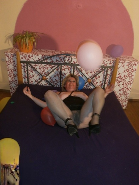 In the midst of balloons on her bed, Caro, an elderly woman, exposes her breast tissue in pantyhose.