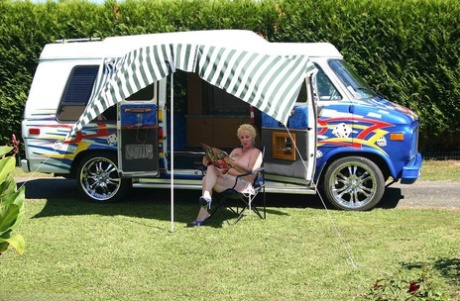 Mary Bitch, a mature blonde woman with big tits and pussy, poses for her camera outside a B class camper.