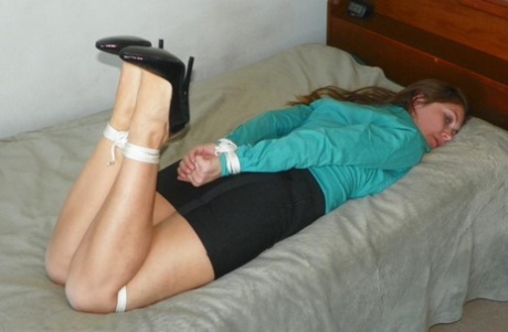 A woman with a broken leg is tied up on a bed, dressed in a black miniskirt and pumps.