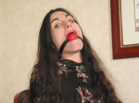 In a photo, the solo girl with long brunette hair exposes her pussy while being restrained and gagged.