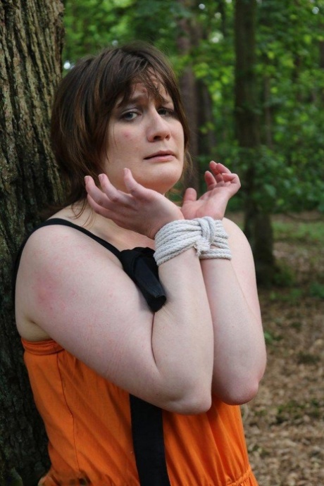 A fat woman is tied loose and pinned to a tree in the woods with her clothes on.