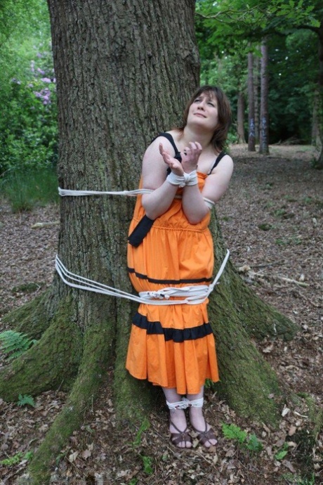 She is gagged and tied to the trunk of a tree in woodland with her garments draped over them.