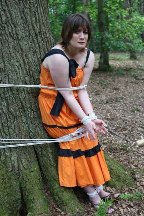 A woman who is overweight is tied loose and pinned to a tree in the woods with her clothing on.