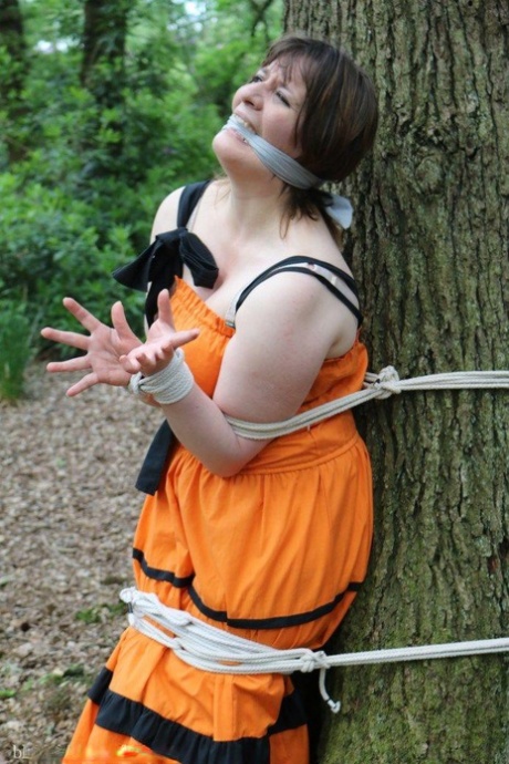 In a forest, an obese woman is tied to a tree and has her clothing put on.