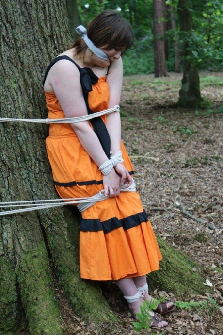 An obese female is bedecked by a tree in the woods and her clothes are tied up, leaving her naked.