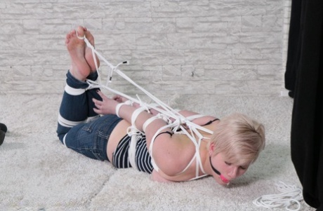 Despite the ball gag and rope bindings, an attractive white girl struggles with her feet.