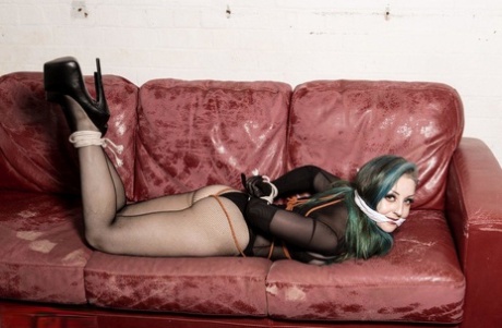 A white girl with dyed hair is gagged and hogtied on a Chesterfield.