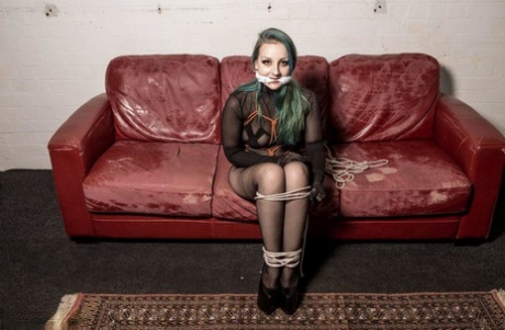 A white girl with dyed hair is gagged and hogtied on a Chesterfield.