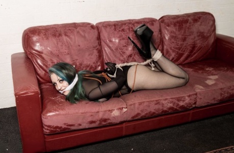 A white girl with dyed hair is gagged and hogtied on a Chesterfield.