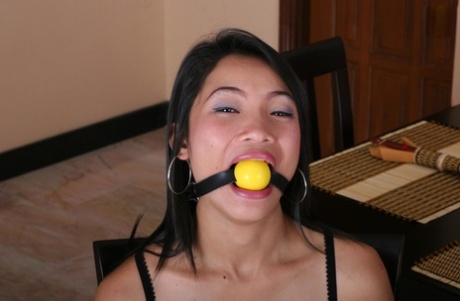 As an Asian girl performs a ball gag while being held to a kitchen chair, she struggles to keep her balance.