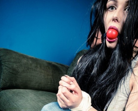 A dark-haired girl performs a big ball gag while being restrained at her wrists and ankles.