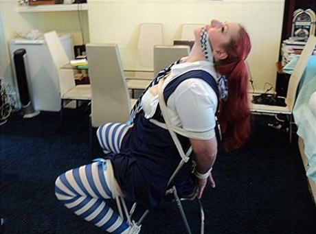 Weighed down, a schoolgirl with heavy red hair is resting on a chair while being gagged with her cleaves.